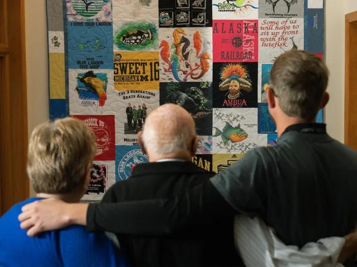 Three people stand arm in arm with backs to camera studying a wall of family mementos