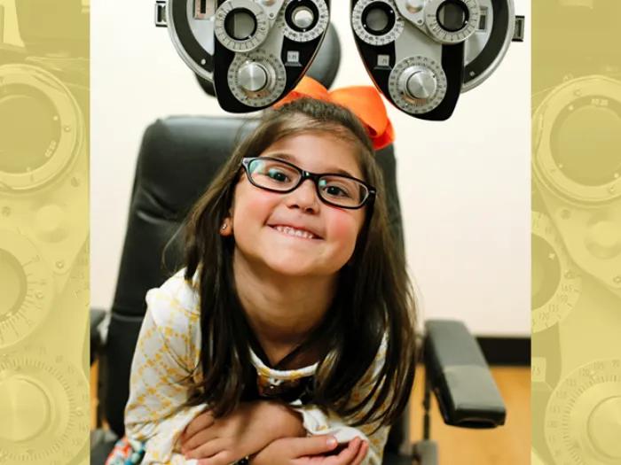 A young girl with glasses smiles while sitting in an eye exam chair under a phoropter.