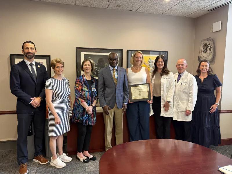Group of eight people of varying ages and races, including Michigan Medicine's Dr. Diane Harper, fifth person from the left. Dr. Harper is a tall white woman, wearing a white blouse and black pants, holding an honorary plaque. . 