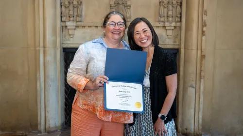 Two women smiling while one holds a certificate from the University of Michigan Medical School, posing together in a formal setting