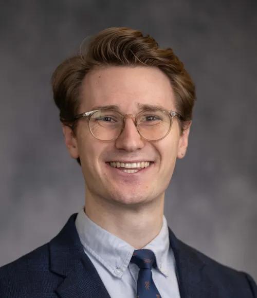 White male with dark red hair and glasses, wearing a dark suit jacket, blue shirt, and dark tie.