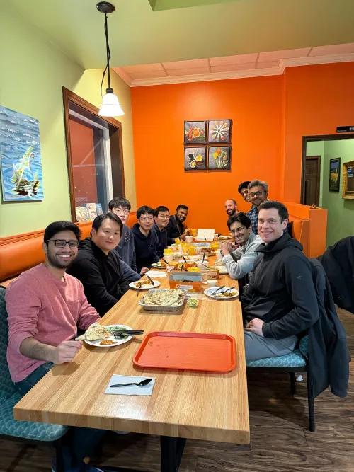 Group seated along a long table in a brightly colored restaurant (orange and green walls), sharing a meal with plates and takeout containers spread across the table.