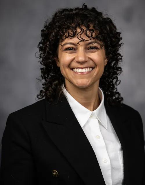 A smiling person with curly dark hair poses for a professional headshot, wearing a black blazer over a white collared shirt against a gray studio backdrop.