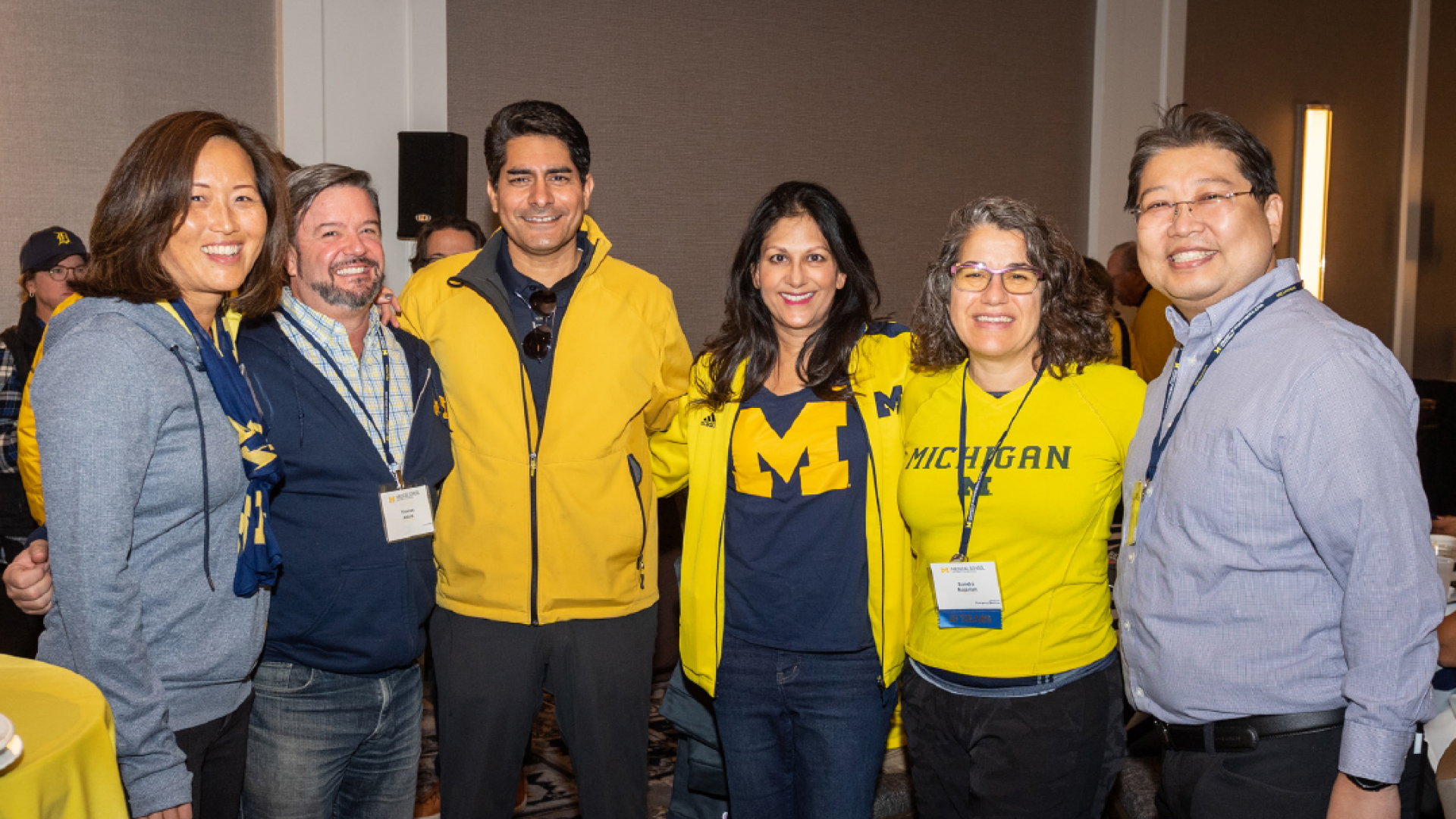 A group of 6 Medical School alumni wearing University of Michigan gear, pose together.