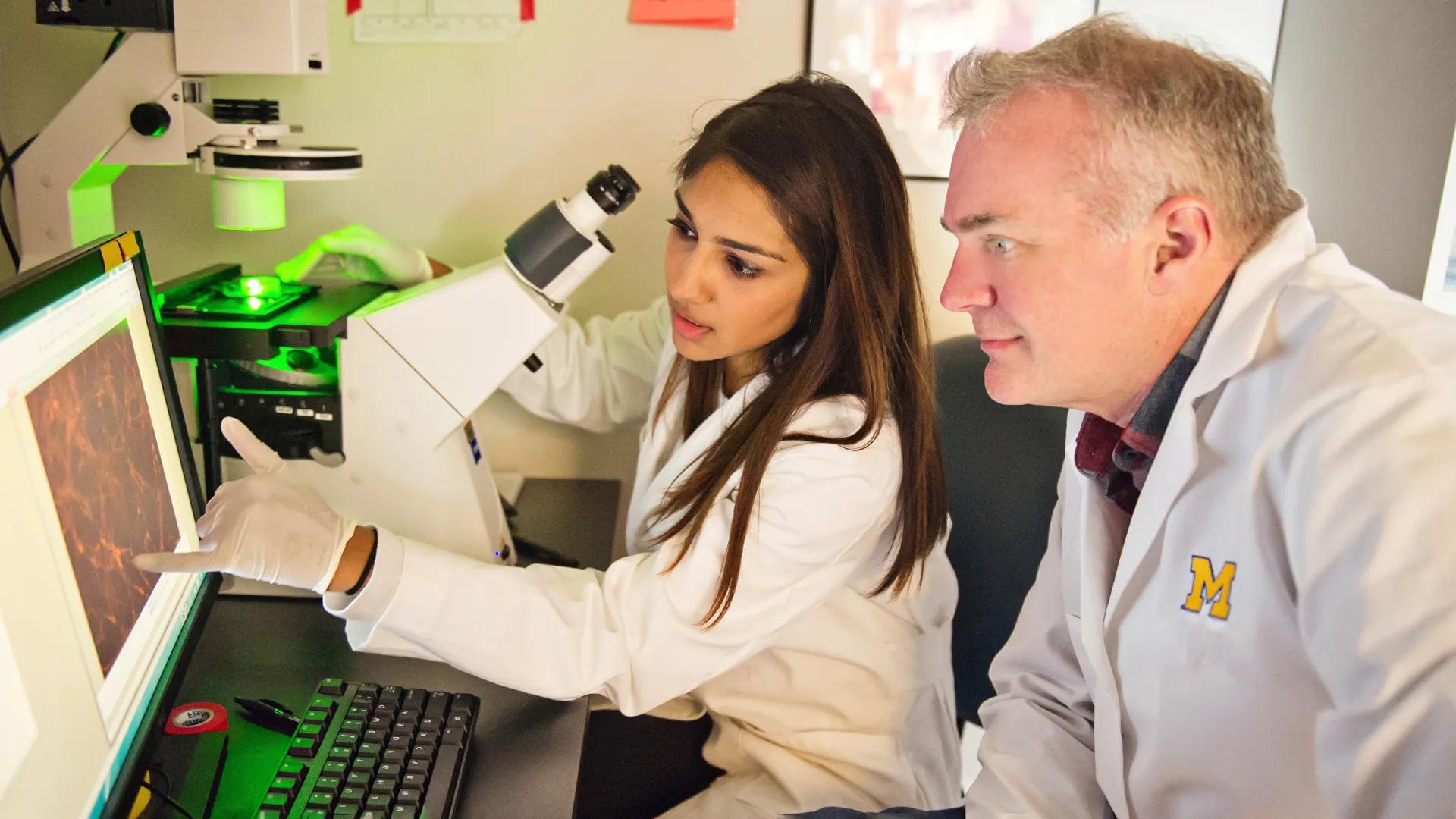 White male and brown-skinned female with long hair wearing white lab coats looking at computer screen with microscope in background