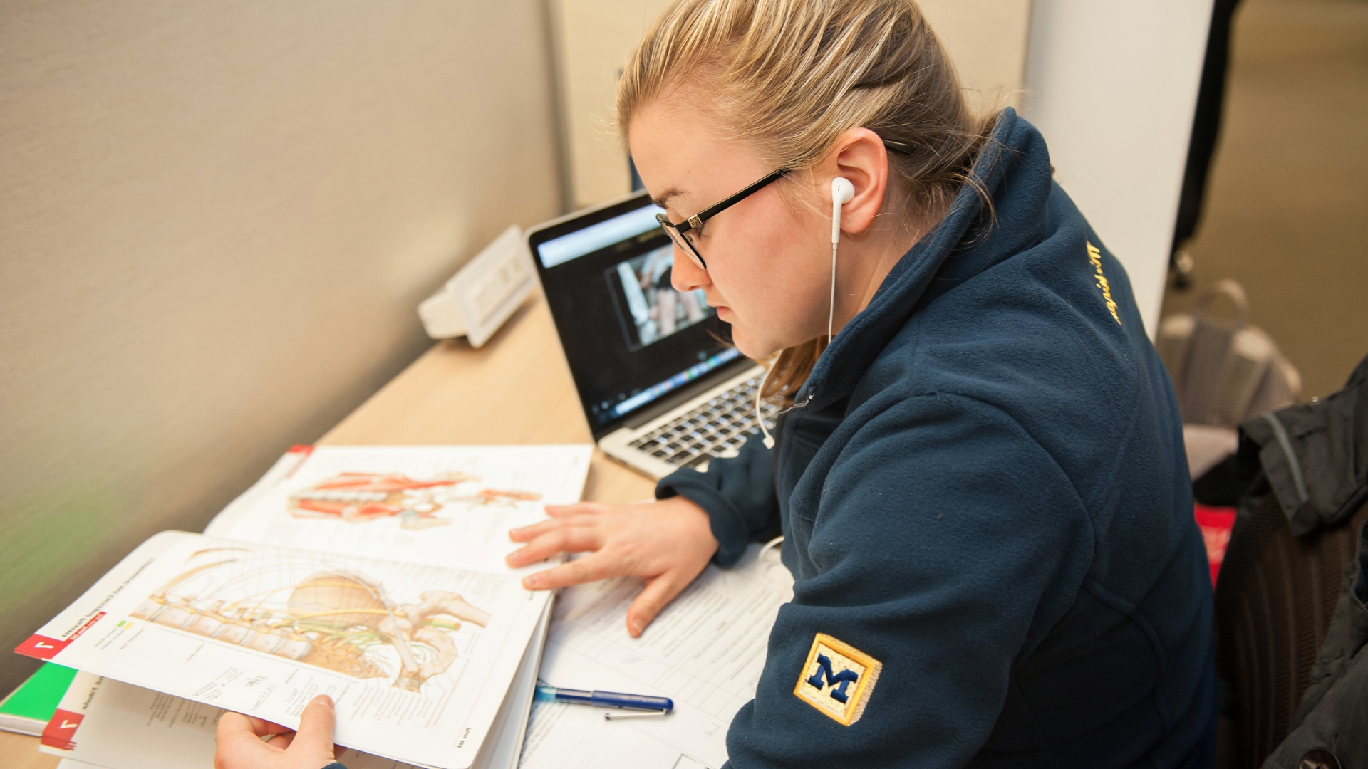 Female student wearing earbuds and Michigan hoodie looking at anatomy textbook with laptop in background