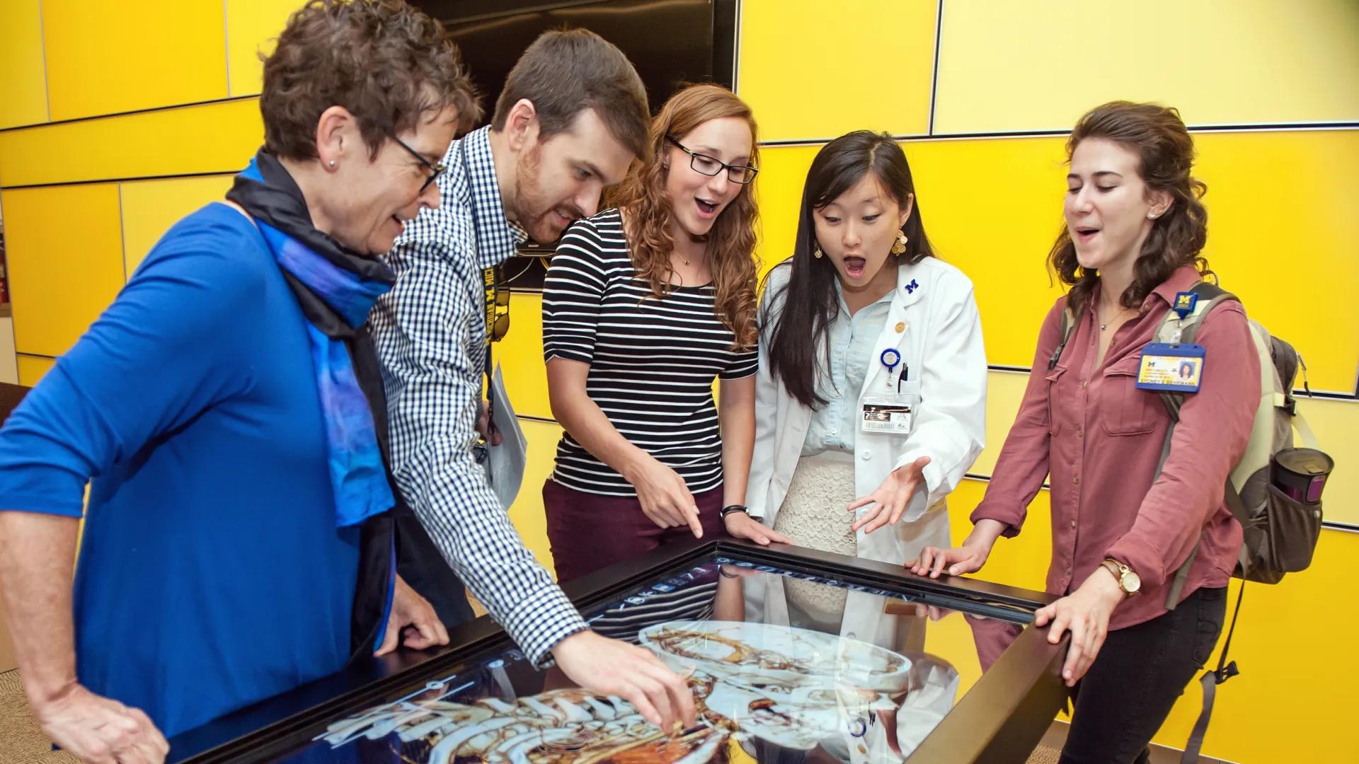 Group of young learners and older woman in blue dress pointing and exclaiming at a medical exhibit in a glass case
