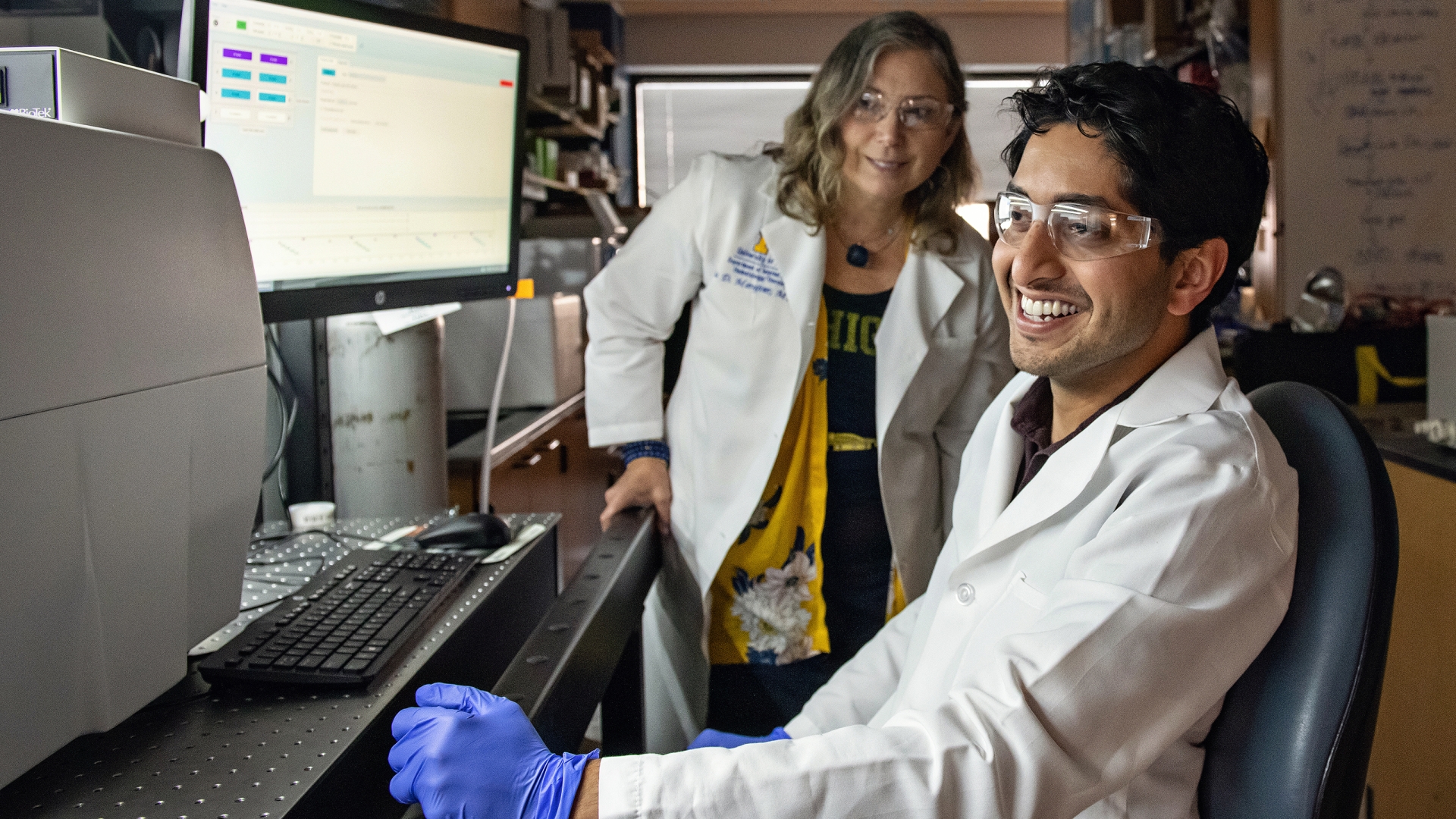 Seated brown-skinned male wearing white jacket and glasses smiling at computer screen with blonde female in white coat smiling in background