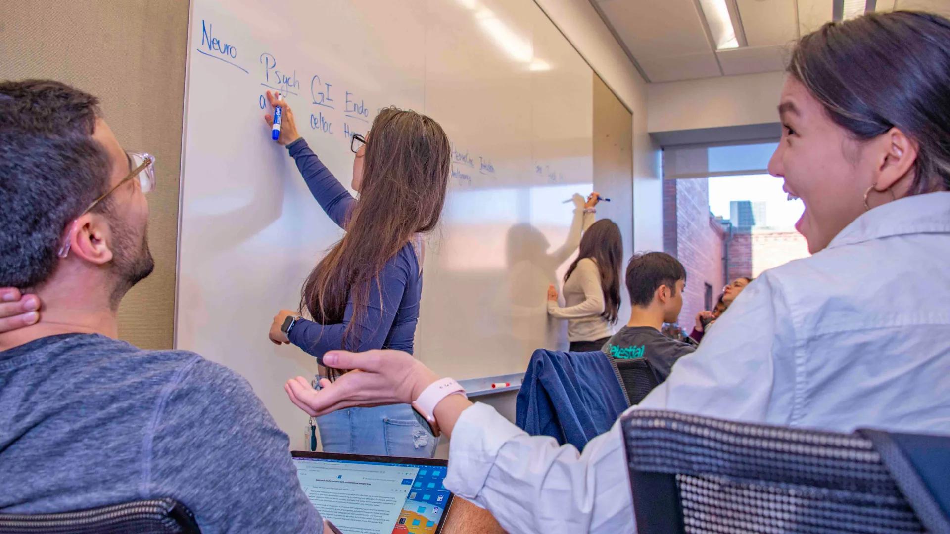 Students laughing together in classroom while others write on a whiteboard