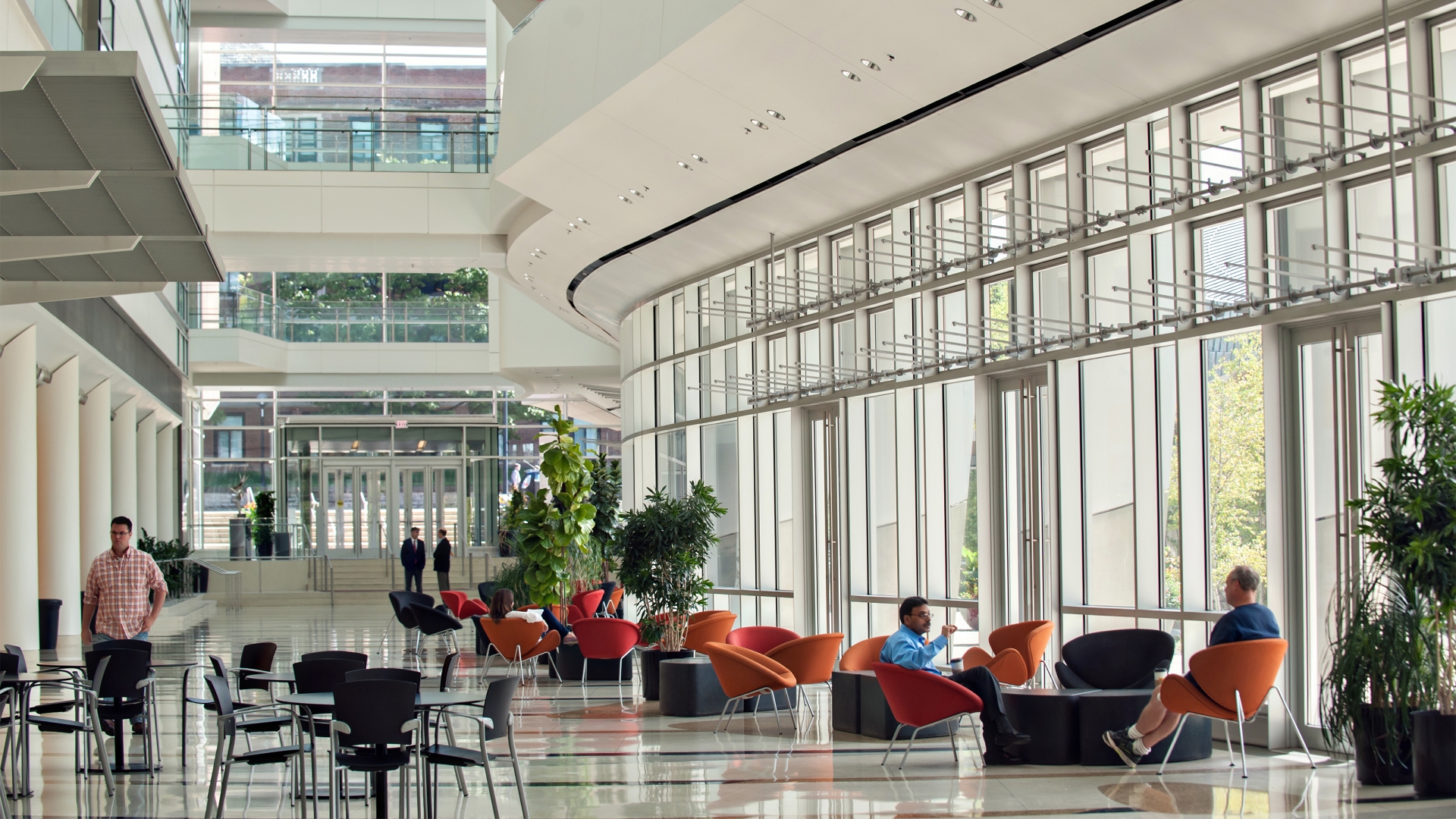 People walking and seated in orange bucket chairs of large modern lobby with curved tall glass walls