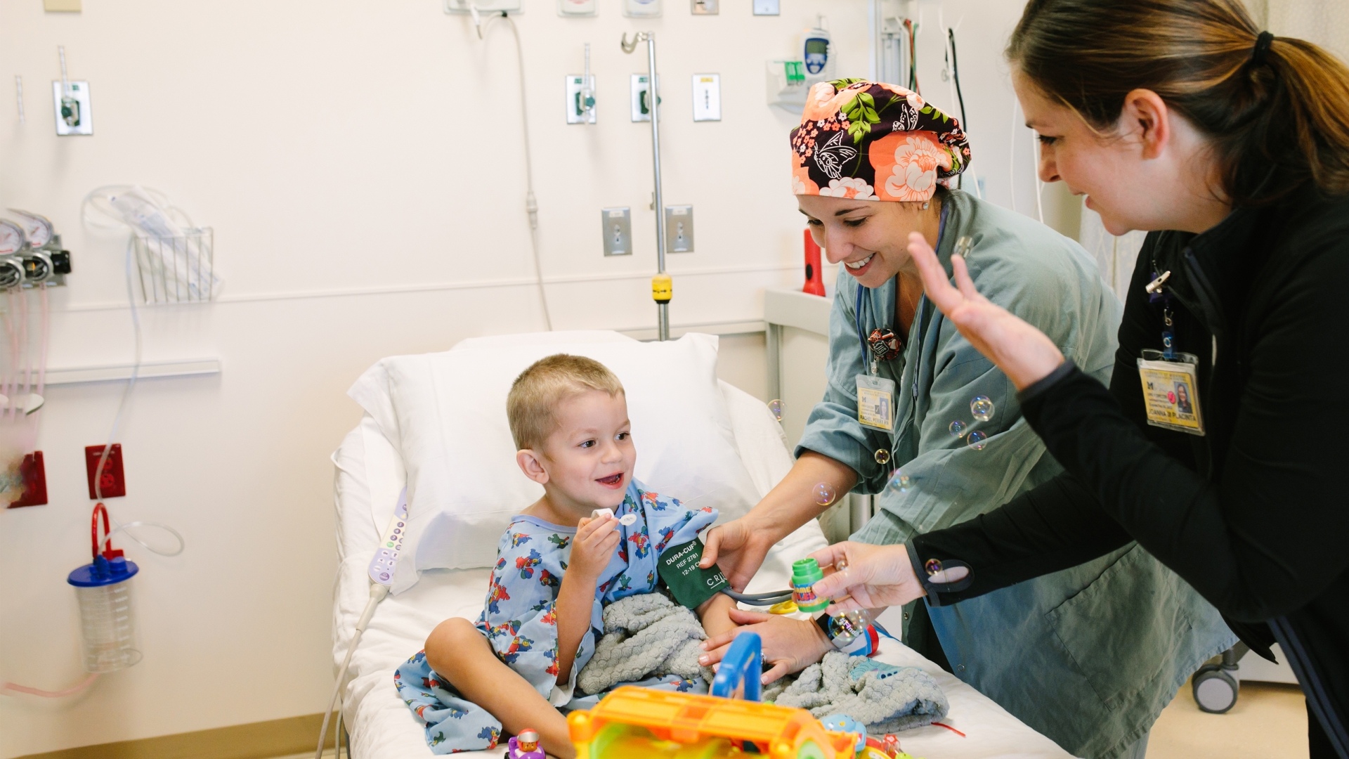 Little boy wearing children's hospital gown in hospital bed with toys and two female medical professionals engaging with him