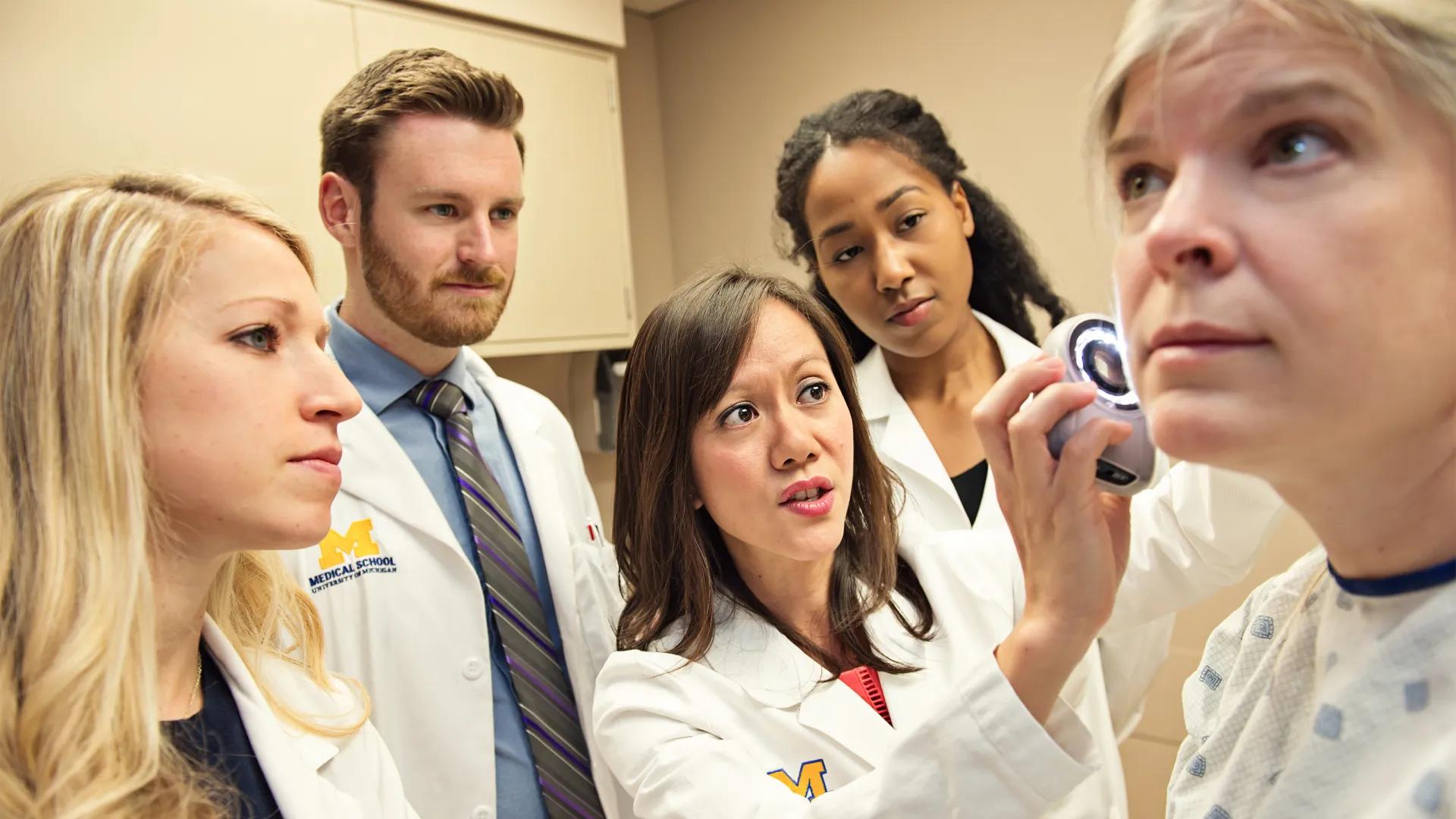 Group of doctors in white coat looking on as one doctor holds an instrument up to a patient's face