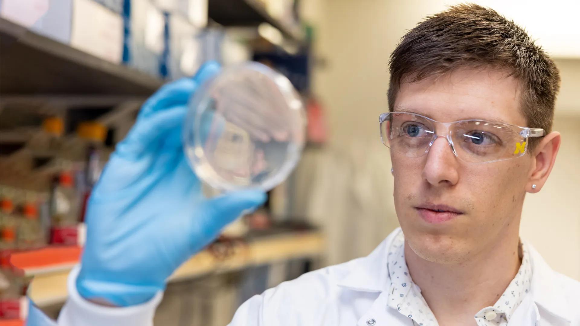 White male with brown hair in lab coat, goggles and gloves holding petri dish