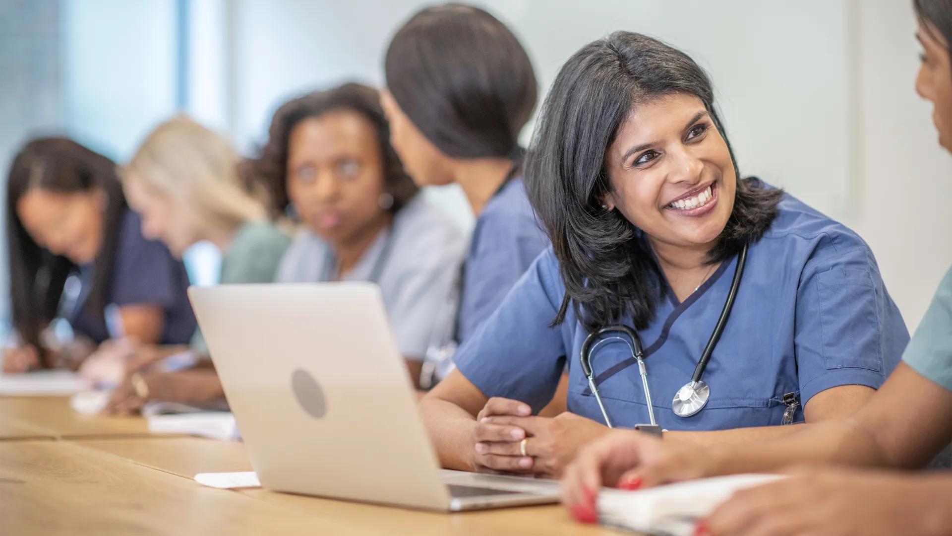 Woman in blue scrubs and stethoscope with laptop smiling at fellow student
