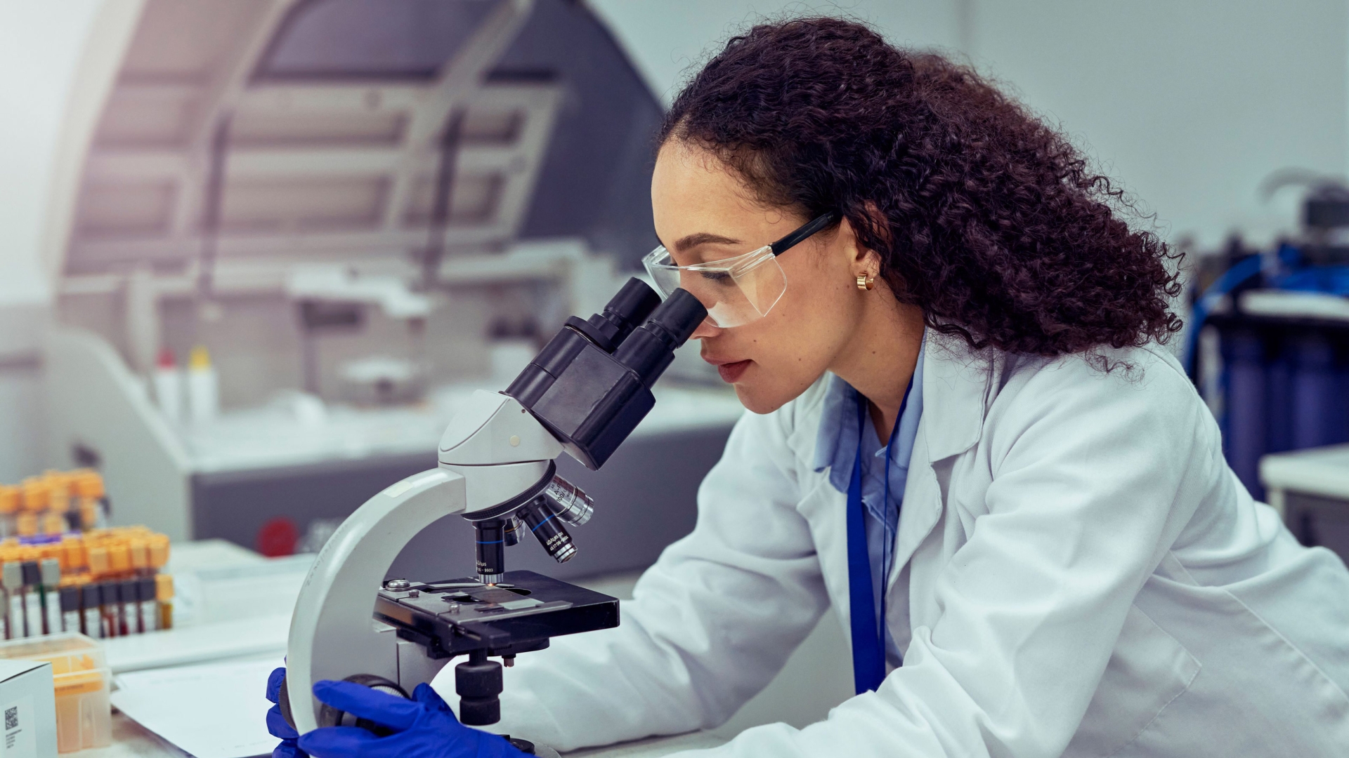 A scientist in a laboratory closely examines a sample under a microscope, wearing protective gloves, a lab coat, and safety goggles.