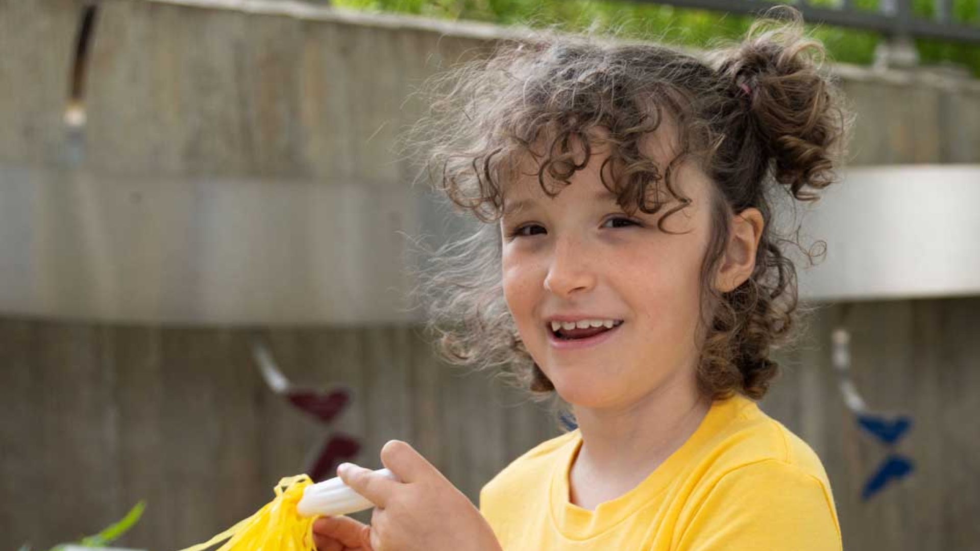 A young girl in a yellow T-shirt with a yellow pompon. She’s outdoors and smiling at the camera. 