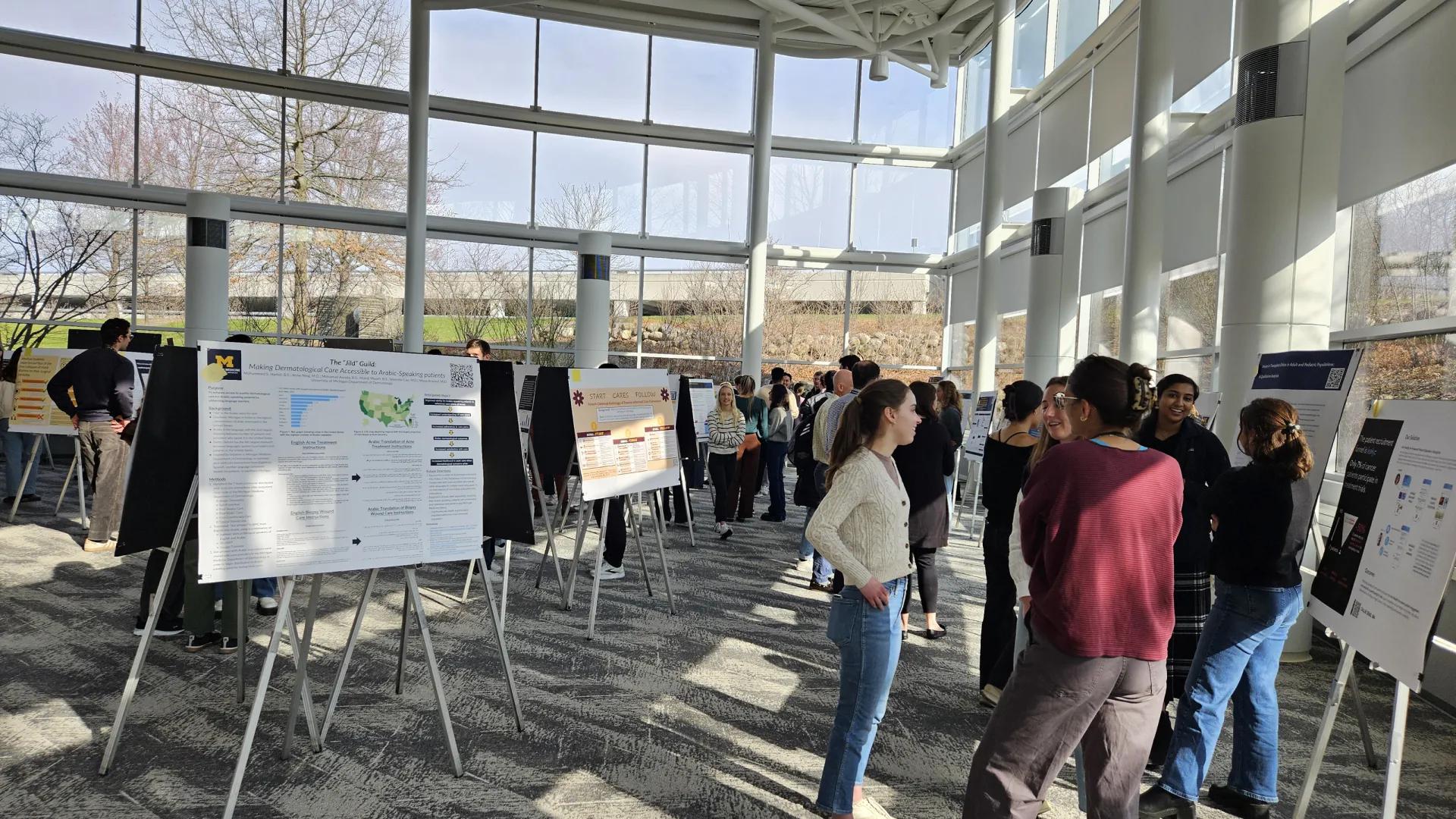 A conference space featuring multiple research poster displays and people standing in groups talking and looking at displays