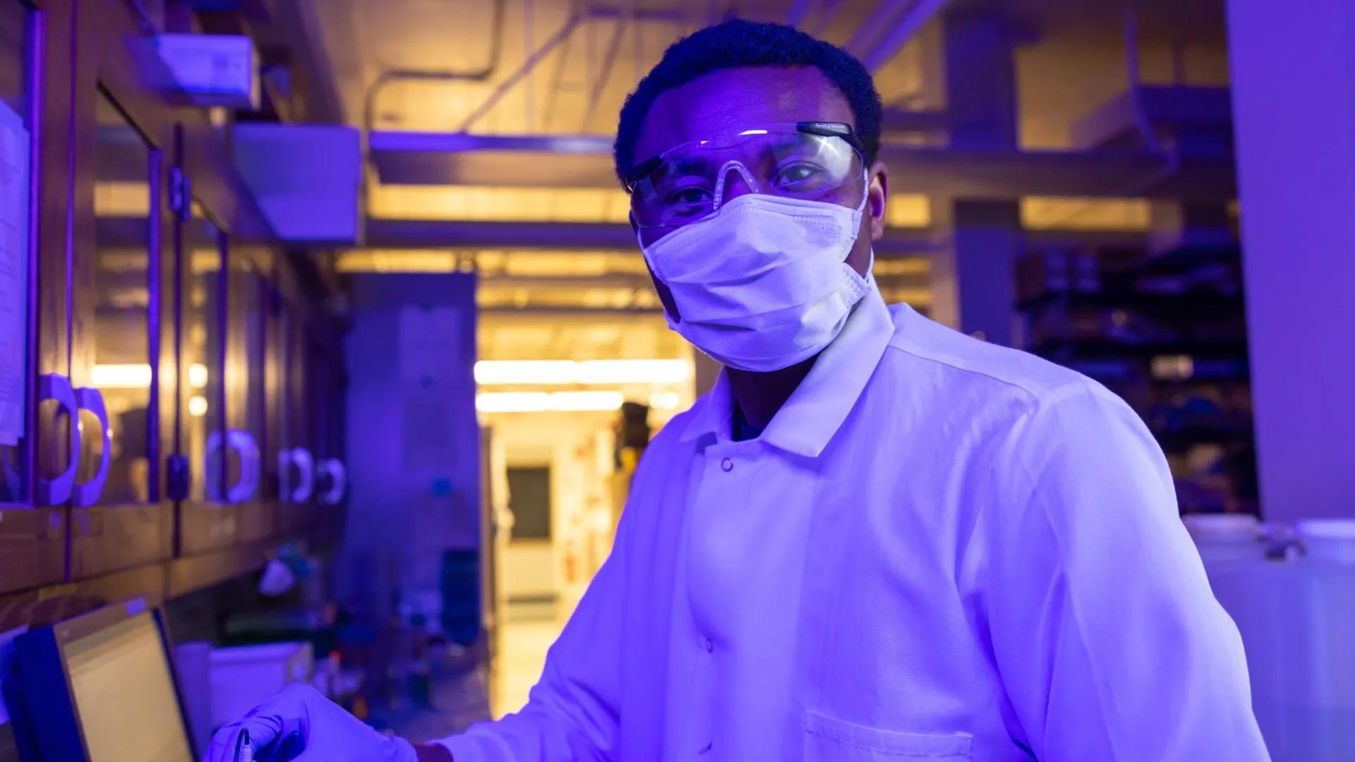 A researcher wearing a mask, goggles, and a lab coat working under blue lighting in a laboratory