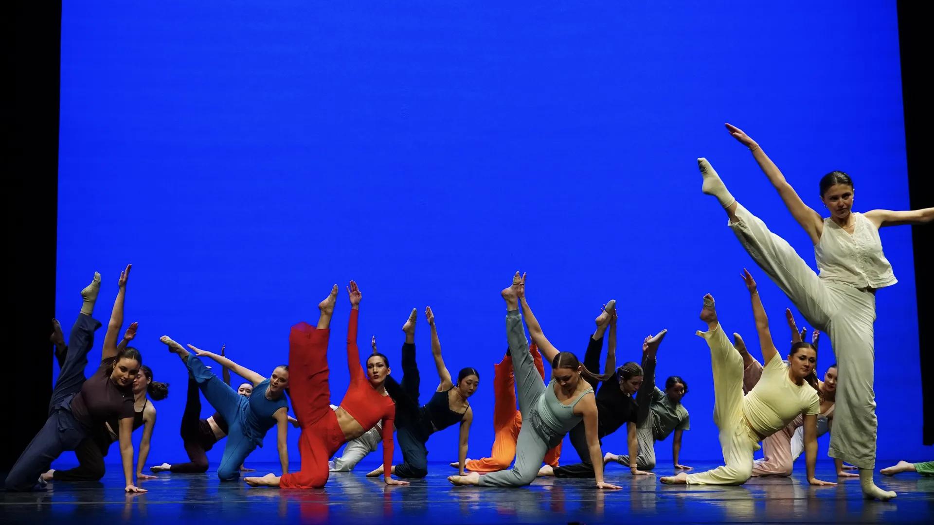 Alt text: A group of dancers performs on a stage with a vivid blue backdrop. Most are low to the floor in a line, balancing on hands with one leg extended vertically, wearing colorful outfits; one dancer at right stands taller, kicking a leg high with arms outstretched.