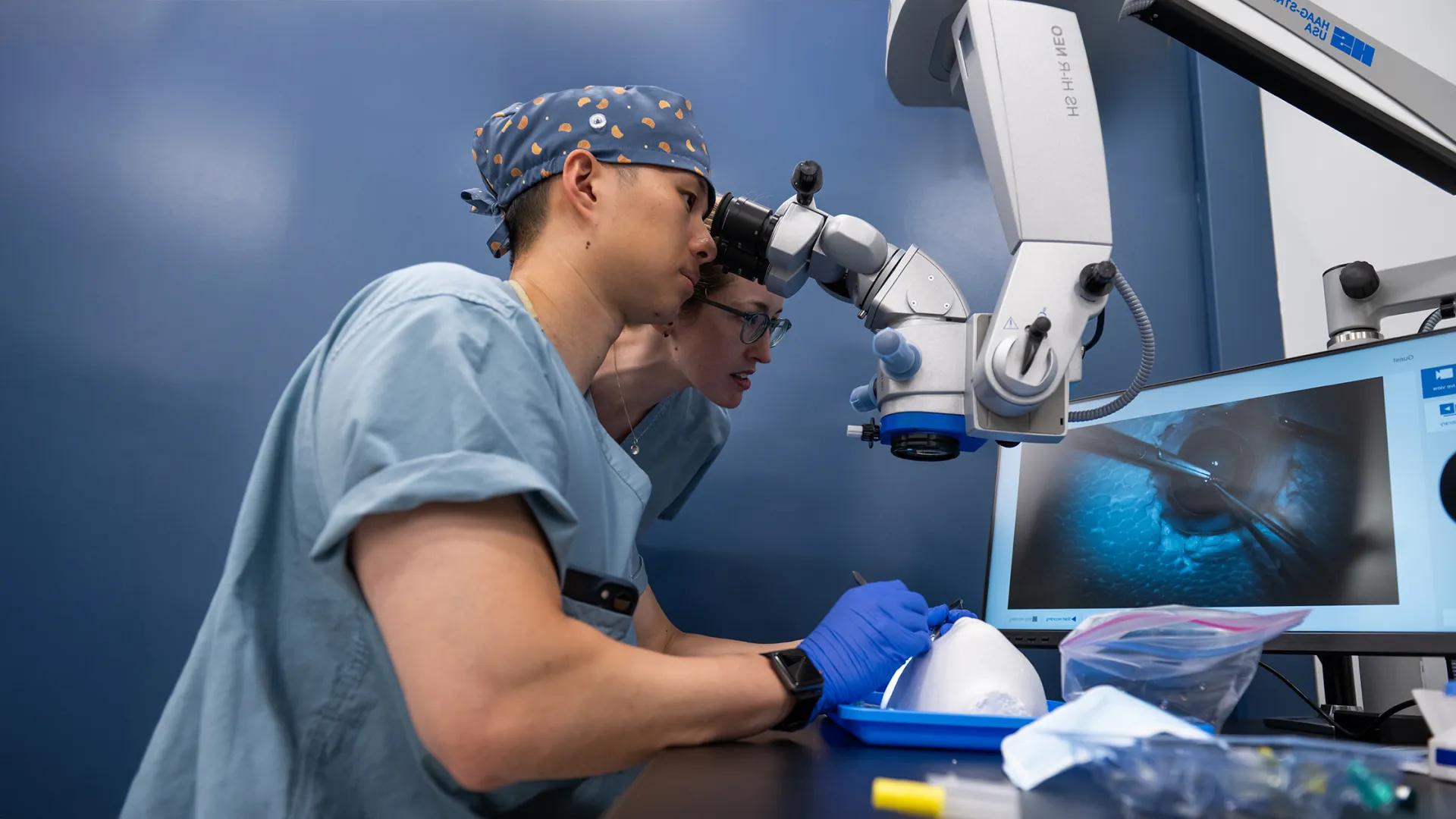 training taking part in microsurgery training with an instructor overseeing. An eye with instruments hovering over it can be seen on a monitor.