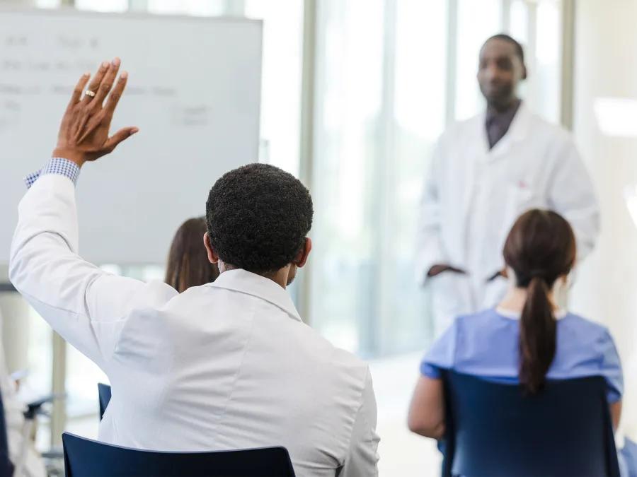 Black doctor standing in front of a group with back showing of student with hand raised