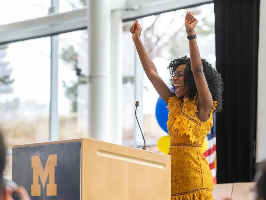 Black woman in yellow lace dress smiling with arms raised in joy behind podium with block M 