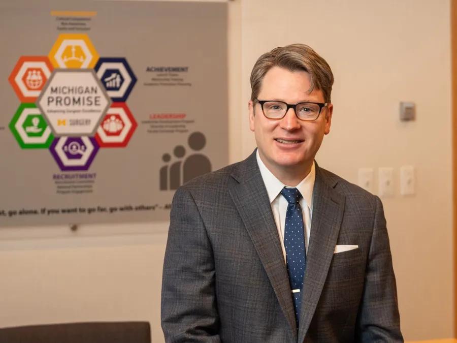 Justin B. Dimick, MD smiles while standing in front of a wall display featuring the Michigan Promise initiative