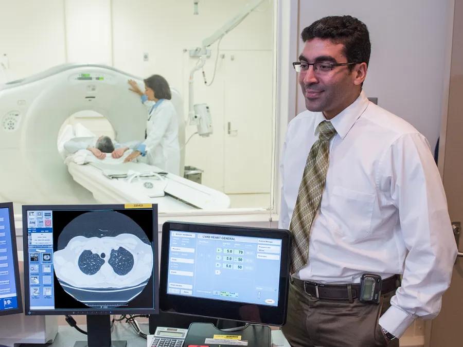 A radiologist standing near CT scan monitors with a patient undergoing a scan in the background