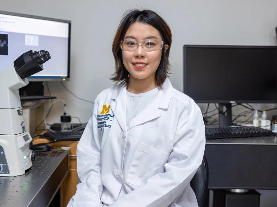 A researcher wearing goggles sits beside a microscope in a laboratory.