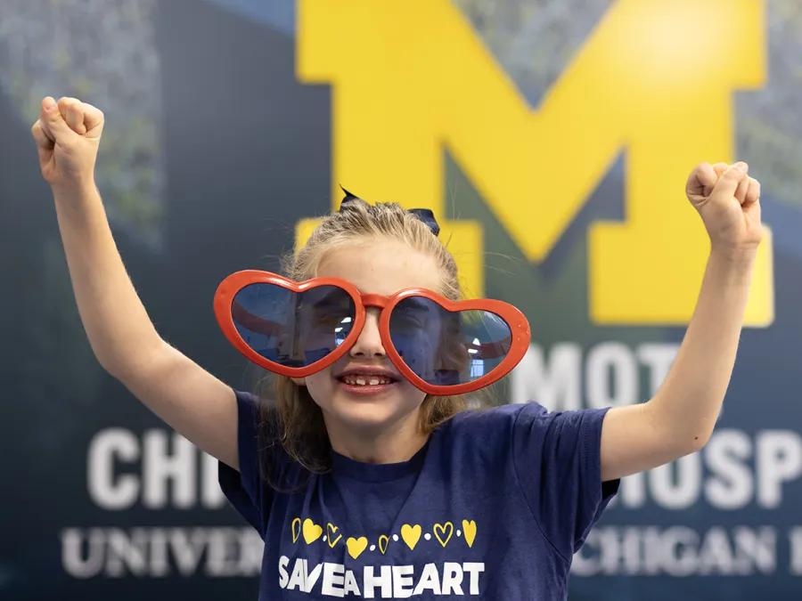 Paisley, a young girl with long brown hair that is pulled back on top, is wearing oversized heart-shaped red sunglasses and a blue Save A Heart t-shirt with yellow hearts across the top. She is standing with her arms extended upwards in a gesture of victory in front of a block M.