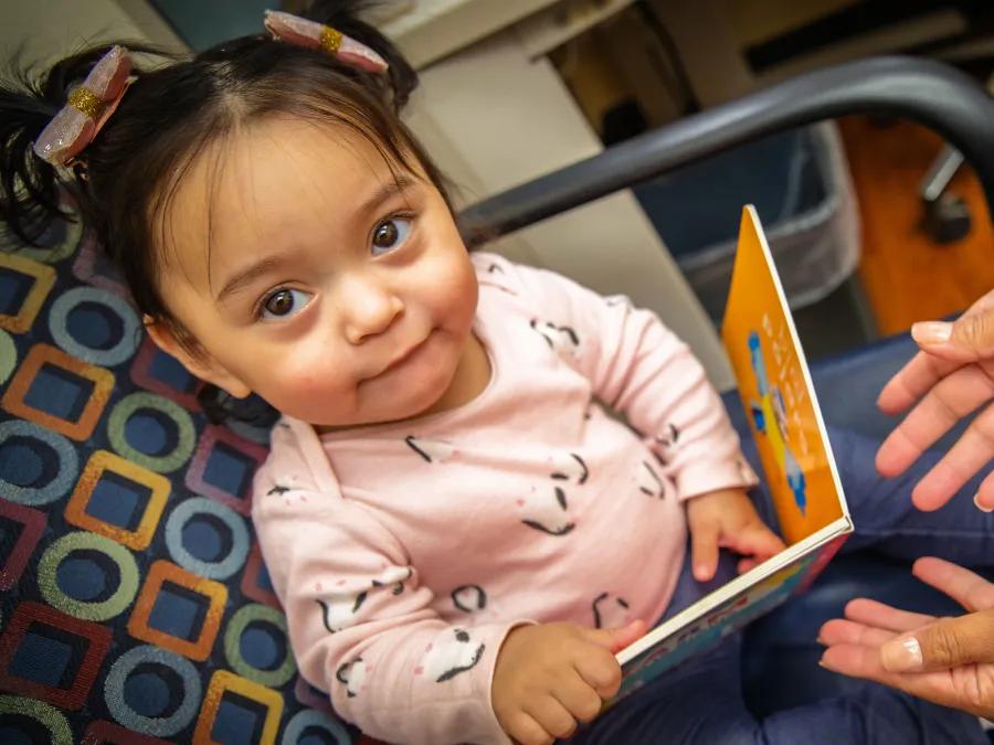 A smiling toddler with pigtails holds a board book while an adult's hands reach out toward them.