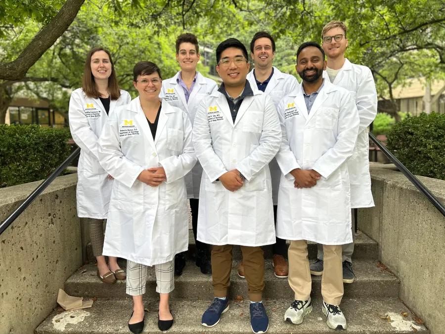 A group of neurology fellows wearing white lab coats standing on outdoor concrete steps with green trees in the background