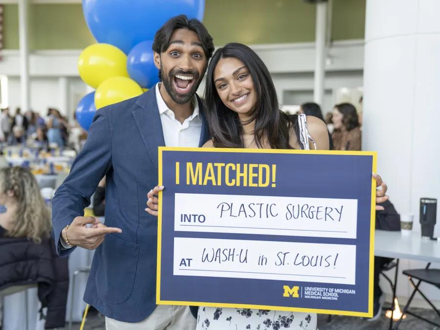 Two people stand together at a celebratory event holding a University of Michigan Medical School sign that reads, “I MATCHED! Into Plastic Surgery at Wash-U in St. Louis!” with blue and yellow balloons and seated guests in the background.