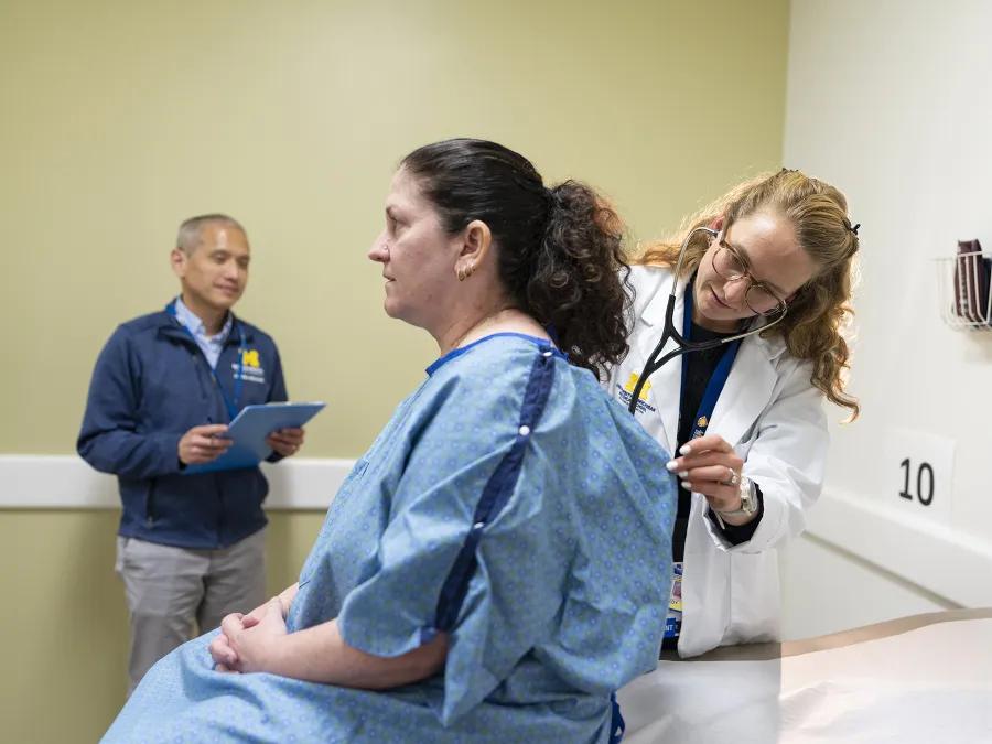 Medical student in a white coat uses a stethoscope to listen to a seated patient’s back in an exam room while a faculty member stands nearby holding a clipboard.