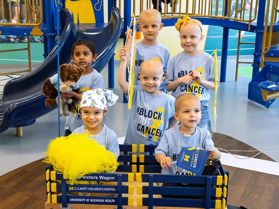 Six kids in Block Out Cancer t-shirts pose by an indoor playground. One holds a Mott teddy bear, two stand in a Mott wagon, and one holds a Maize pom-pom.