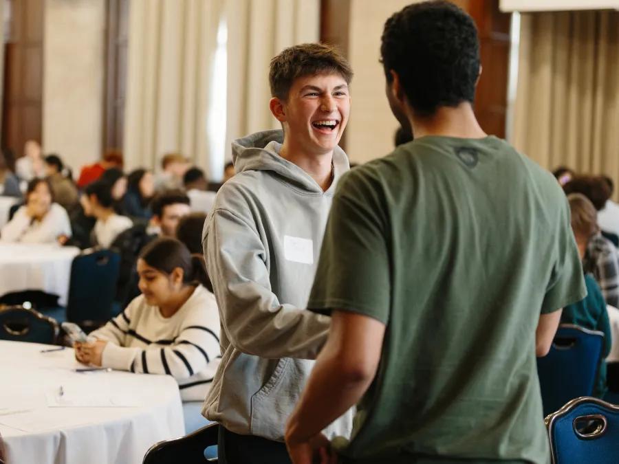 A smiling student in a gray hoodie greets another student in a green T-shirt during a lively group event in a large meeting room.