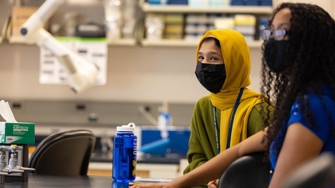 Two women wearing masks, one wearing a gold hijab, the other a Black woman wearing glasses, in a laboratory setting
