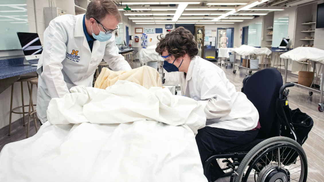 A professor in a white coat and mask discusses anatomy with a medical student in the anatomy lab. They are both leaned over an anatomical donor, who is covered up with a sheet.