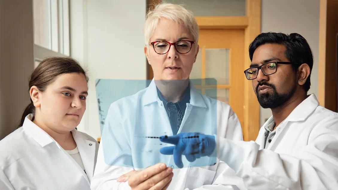 Group of two females and one male wearing white coats and looking at a pale blue transparency 