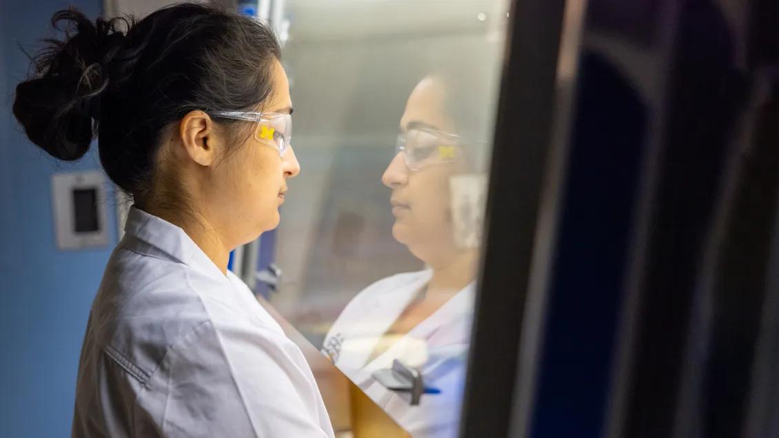 Woman with dark hair in a high ponytail wearing white coat and research goggles with block M reflected in glass
