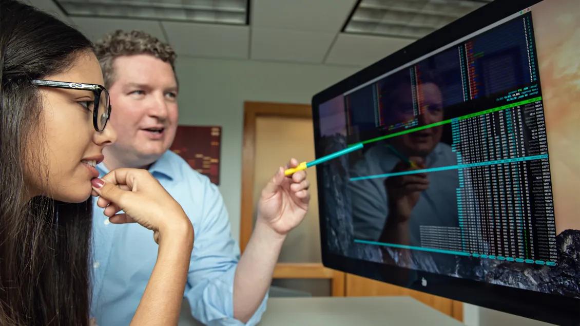 Woman with long dark hair and glasses with man in pale blue shirt pointing at data on large screen
