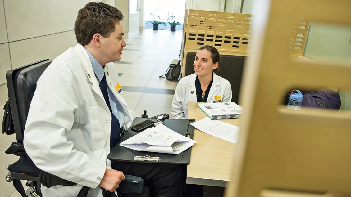 Young Caucasian doctor in wheelchair seated at desk talking to seated young female doctor
