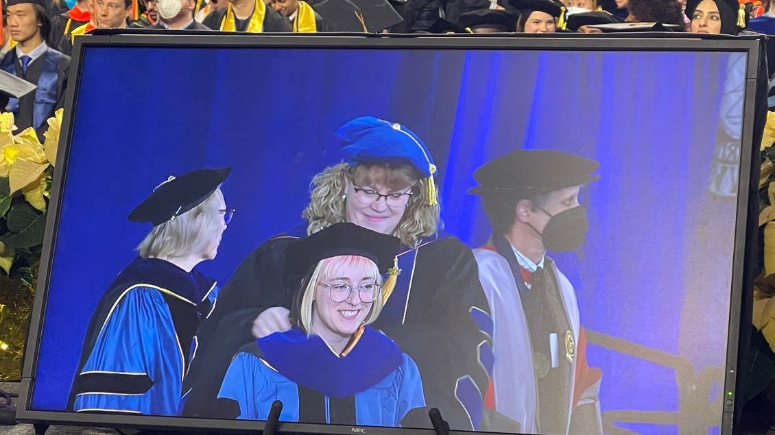 A live event being displayed on a large screen, showing a group of individuals wearing graduation caps and gowns