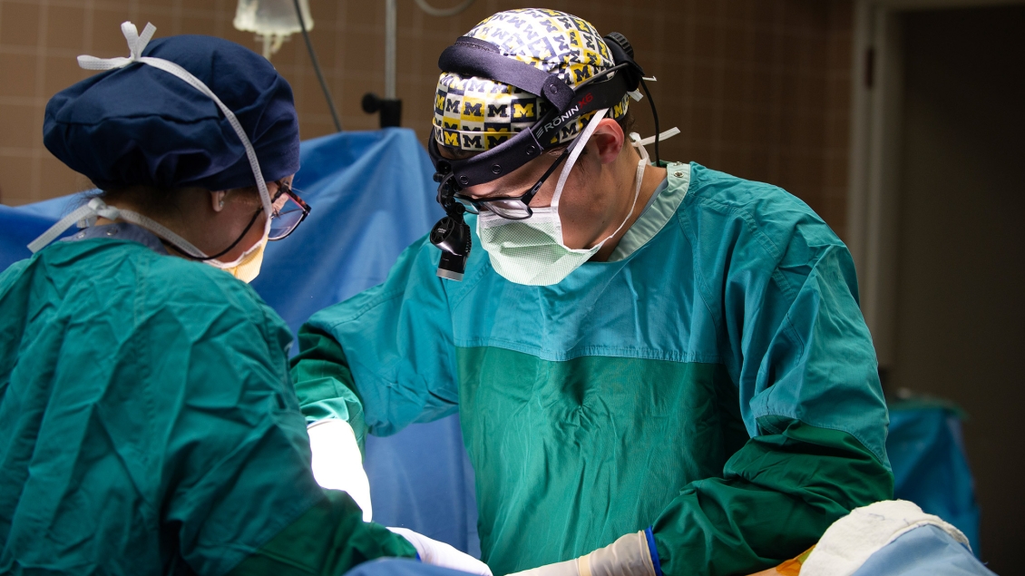 Two surgeons focused on a procedure in an operating room, dressed in green surgical gowns and wearing head coverings and masks, with one surgeon using a head-mounted magnifying loupe.