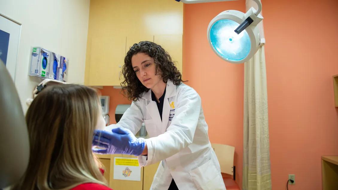 Doctor wearing a white coat examines a patient's face in a medical clinic, using gloved hands under a bright overhead light.