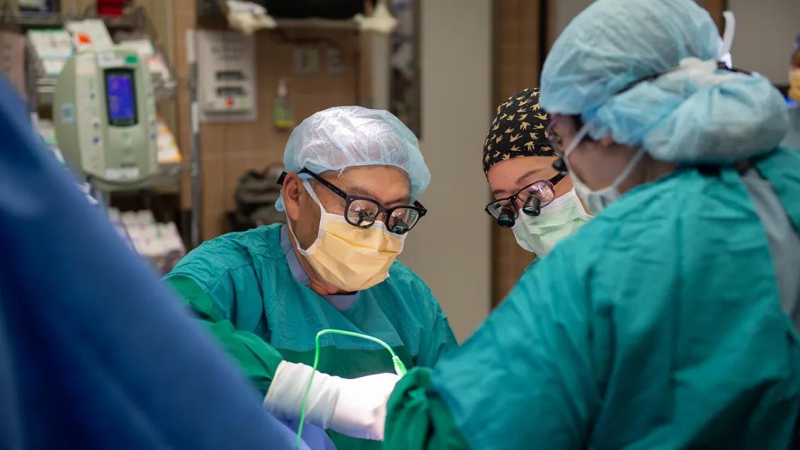  Three surgeons, dressed in surgical gowns, masks, and protective eyewear, are focused on a procedure in an operating room