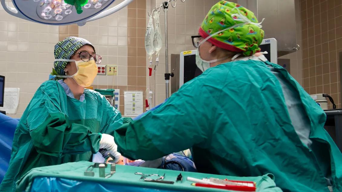  Two surgeons in an operating room, wearing surgical gowns, masks, and colorful head coverings, work together on a procedure under bright surgical lights.