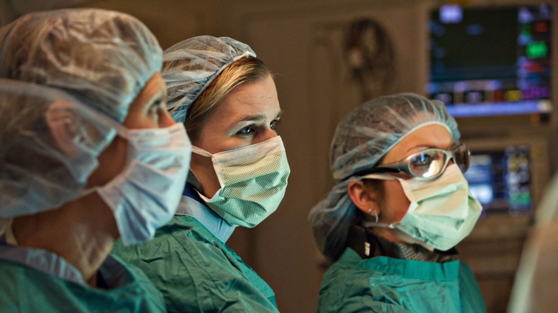 Three surgeons in an operating room observe and assist in a procedure, with medical monitors visible in the background.