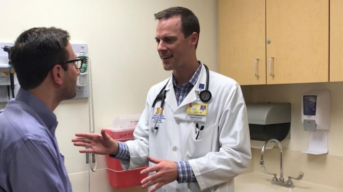 A doctor in a white coat talks with a seated patient in an exam room.