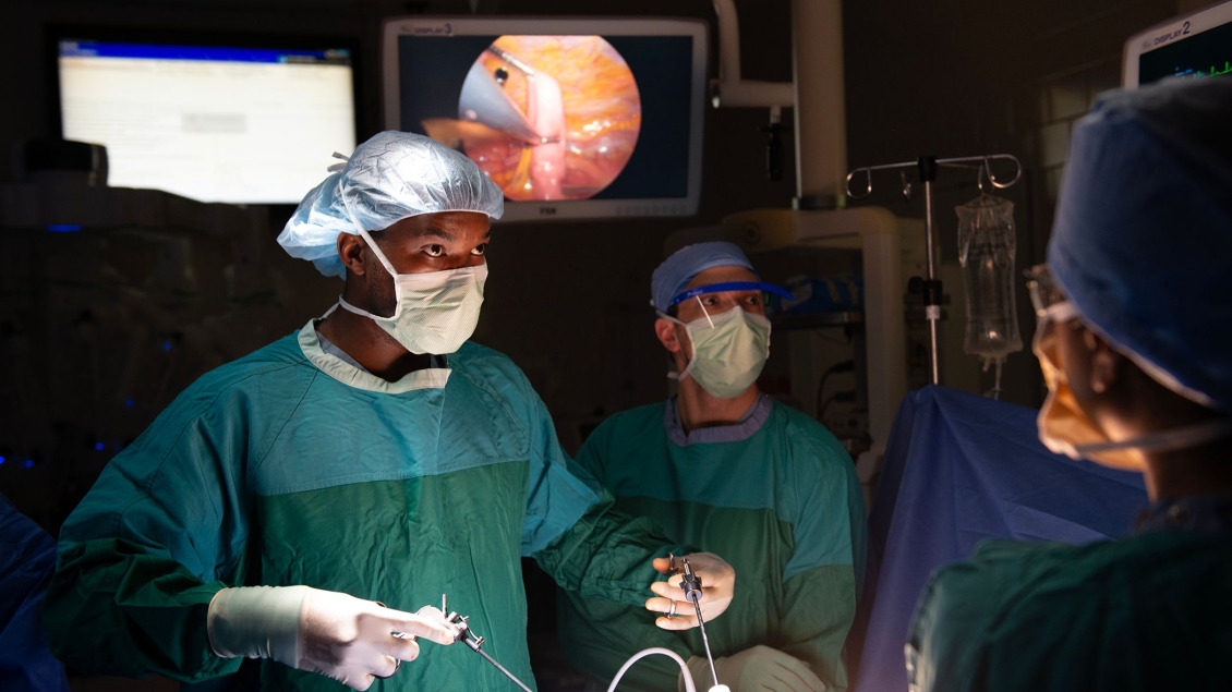 A surgeon maneuvers laparoscopic tools in an operating room as a medical student watches.